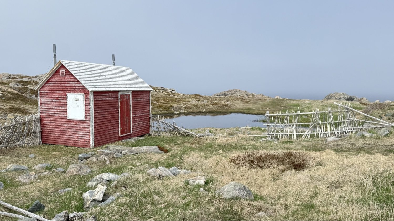 A red fishing shed with a wooden fish-drying flake behind it, near a small pond