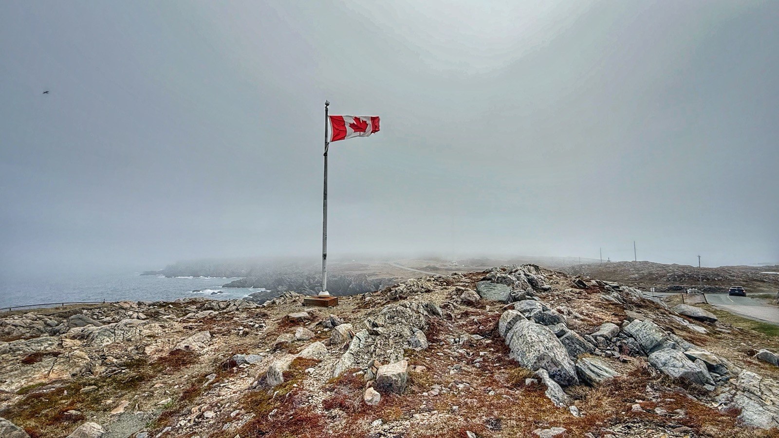 A Canadian flag flying atop a rocky point near the lighthouse, with the foggy coastline stretching into the distance