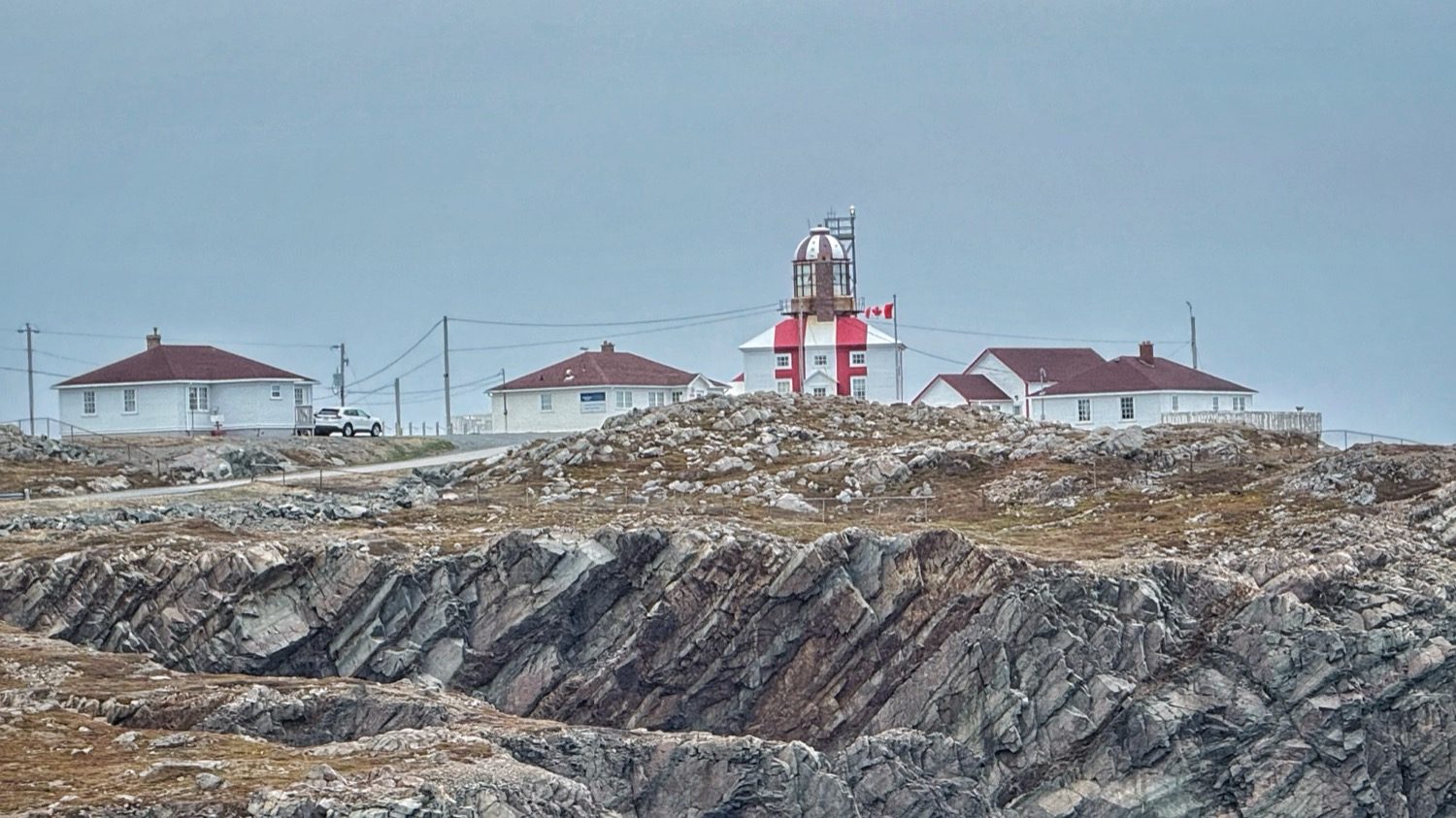 The red and white Cape Bonavista Lighthouse and its outbuildings on a rocky point, seen from a distance