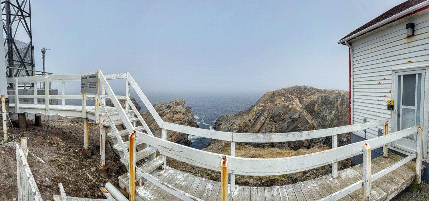 A white wooden walkway with a posted warning sign, connecting the lightkeeper's house to a transmission tower above the cliffs