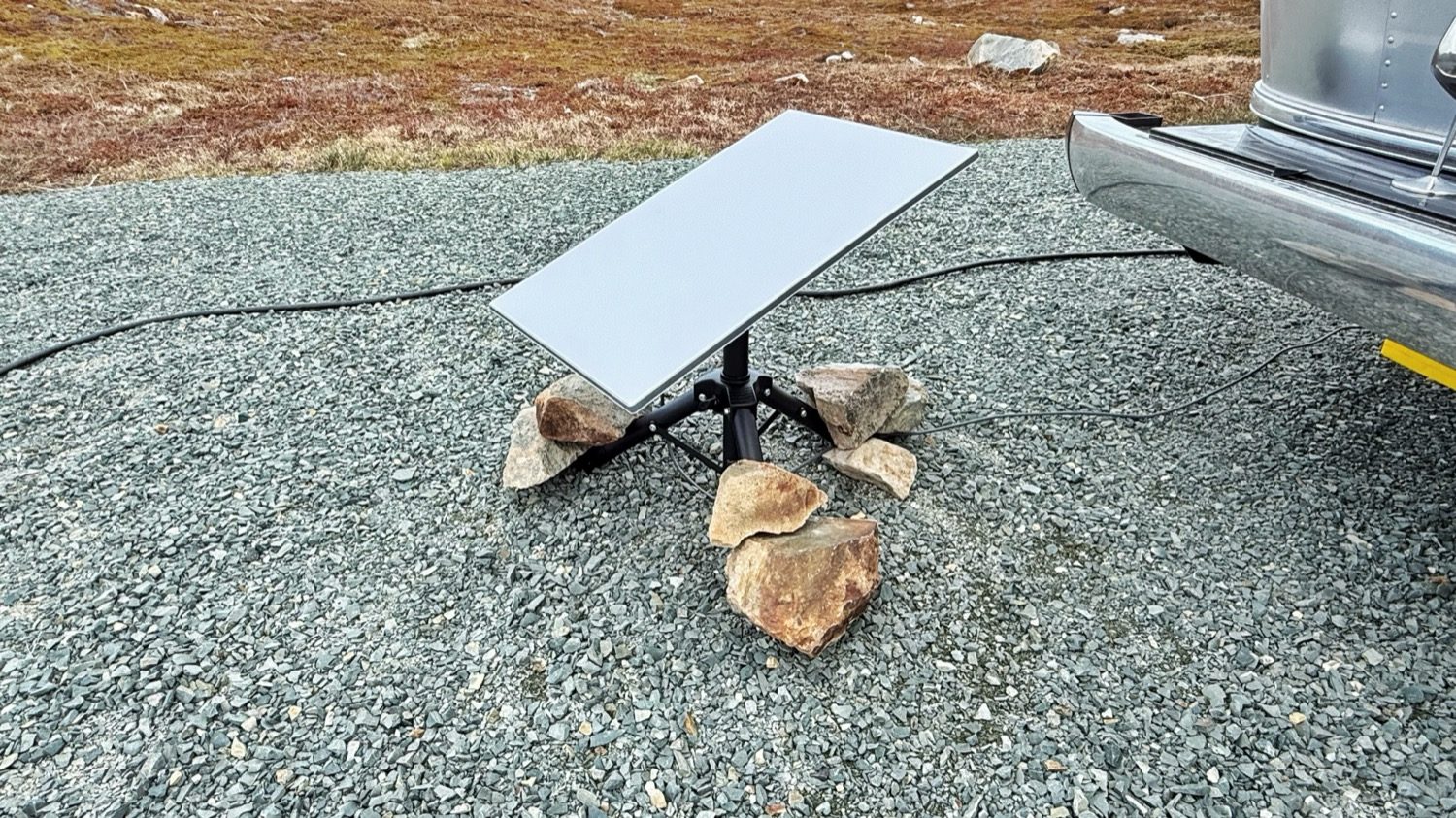 A Starlink satellite dish weighted down with rocks on a gravel lot, with the Airstream behind it