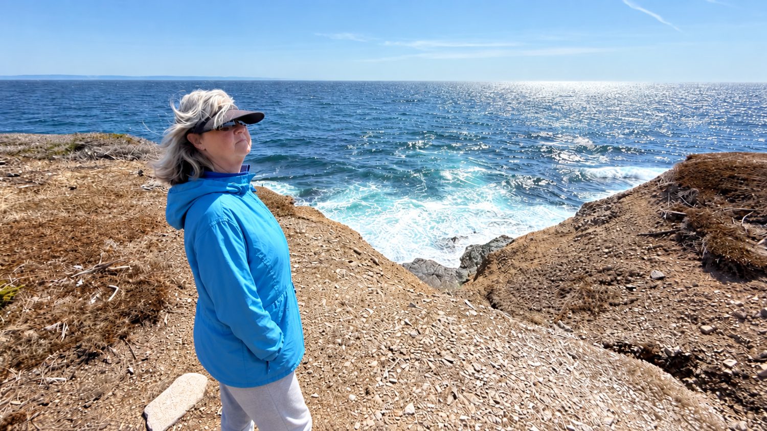 Heather standing near the top of an unguarded cliff at Boutte du Cap Park, on loose dirt and gravel ground, with a steep drop to the water just beside her