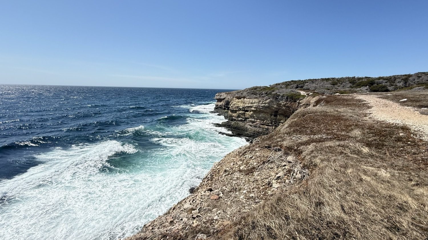 A gravel trail running along the cliff edge at Boutte du Cap Park, with waves breaking on the rocks below