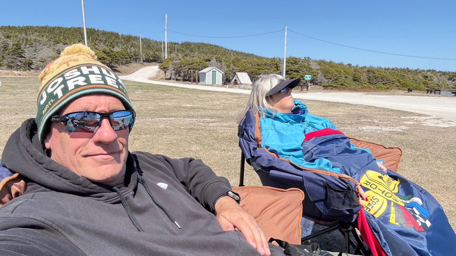 Dan and Heather relaxed in camp chairs at Boutte du Cap Park, out of the wind, with the park grounds and a gravel road behind them