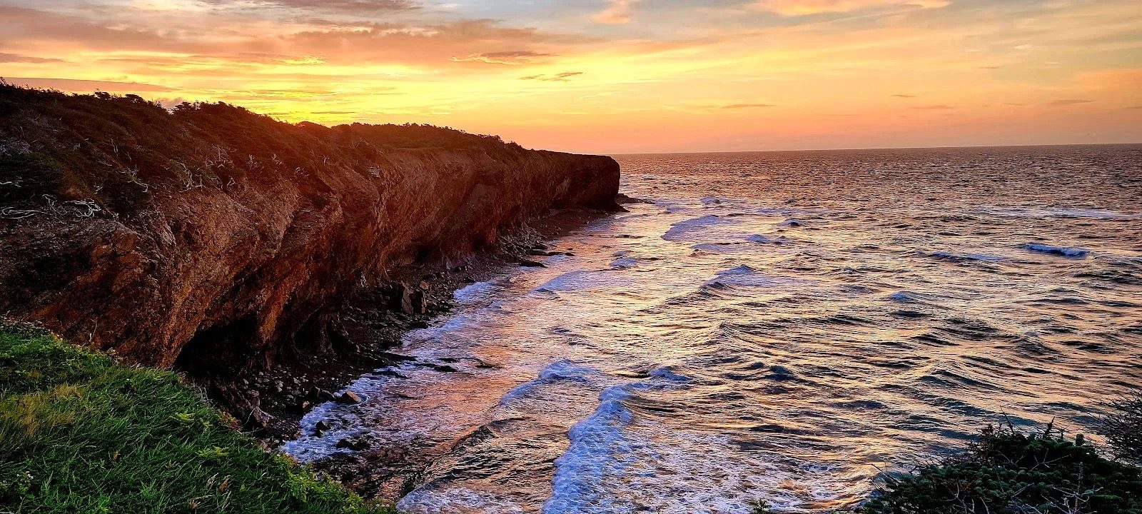 Sunset over the cliffs at Boutte du Cap Park, with orange light on the water and waves breaking below the headland