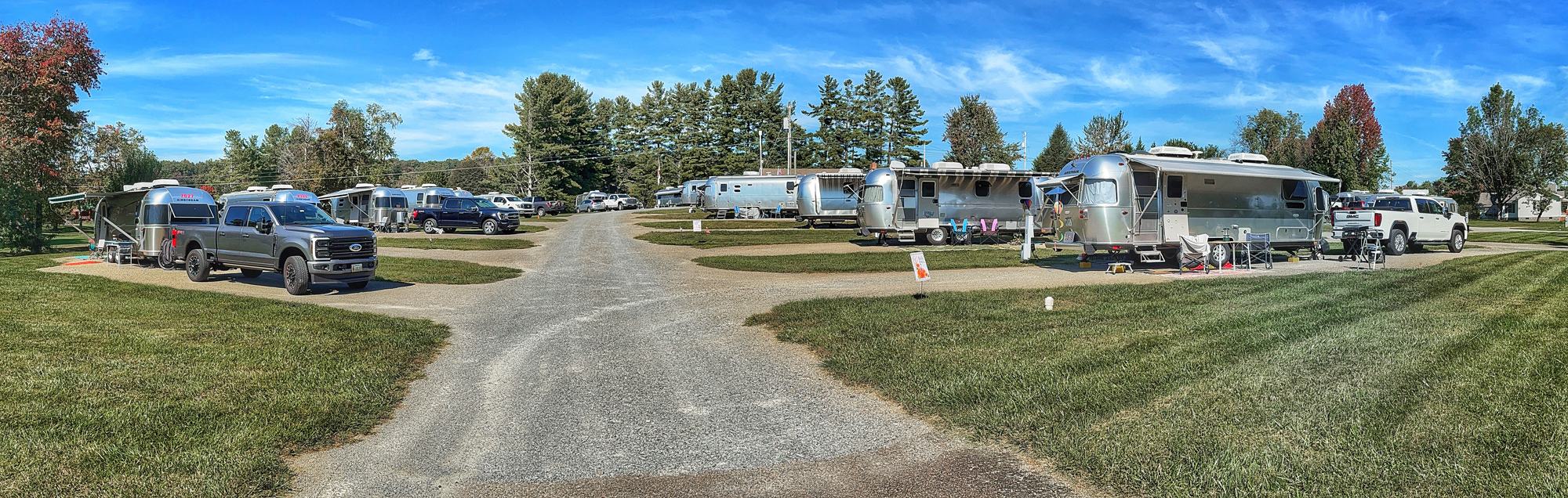 A panoramic view of Airstream trailers and tow vehicles parked on both sides of a gravel lane, under a blue sky with autumn-colored trees behind them