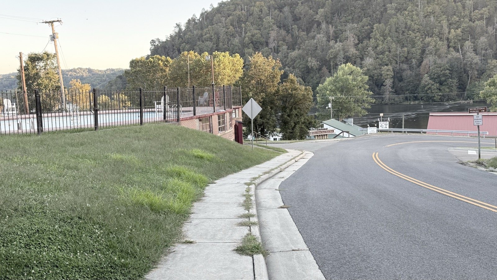 A curving street dropping toward the New River in the early evening, with a brown New River Trail State Park sign at the bottom and forested hills across the water