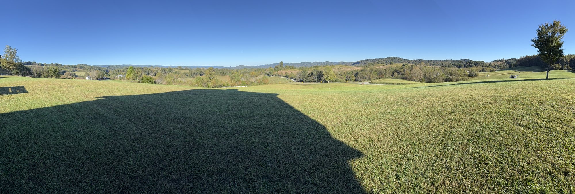 A panoramic view across a mown grassy hilltop toward rolling green hills and distant blue mountains under a clear sky, with a long dark shadow stretching across the grass in the foreground