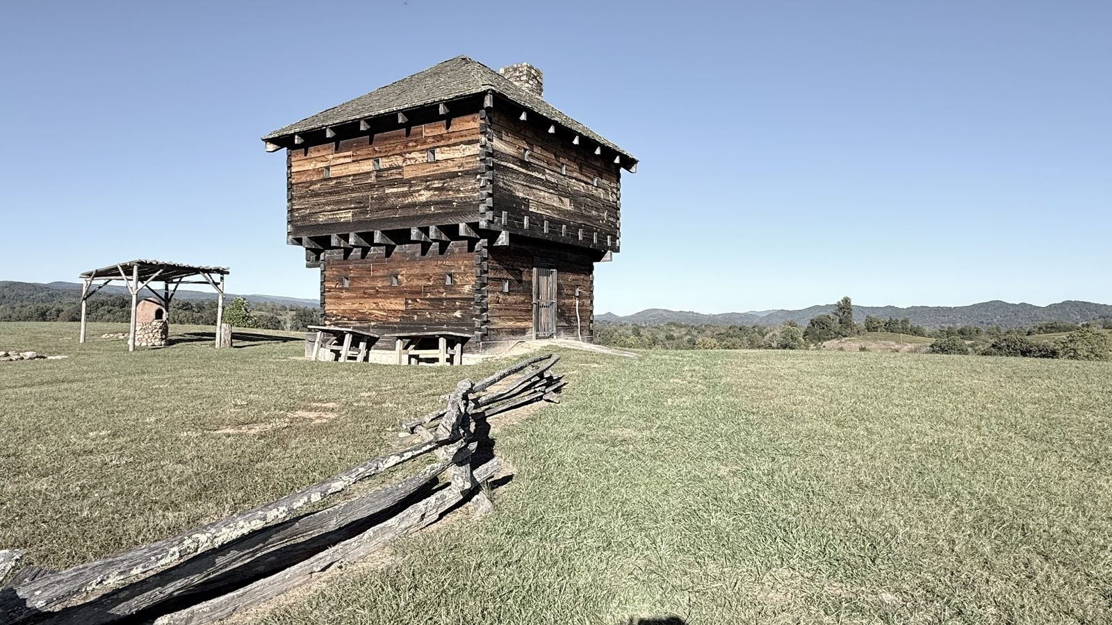A log blockhouse with a hip roof and stone chimney on a grassy hilltop, with a split-rail fence in the foreground and distant mountains behind it