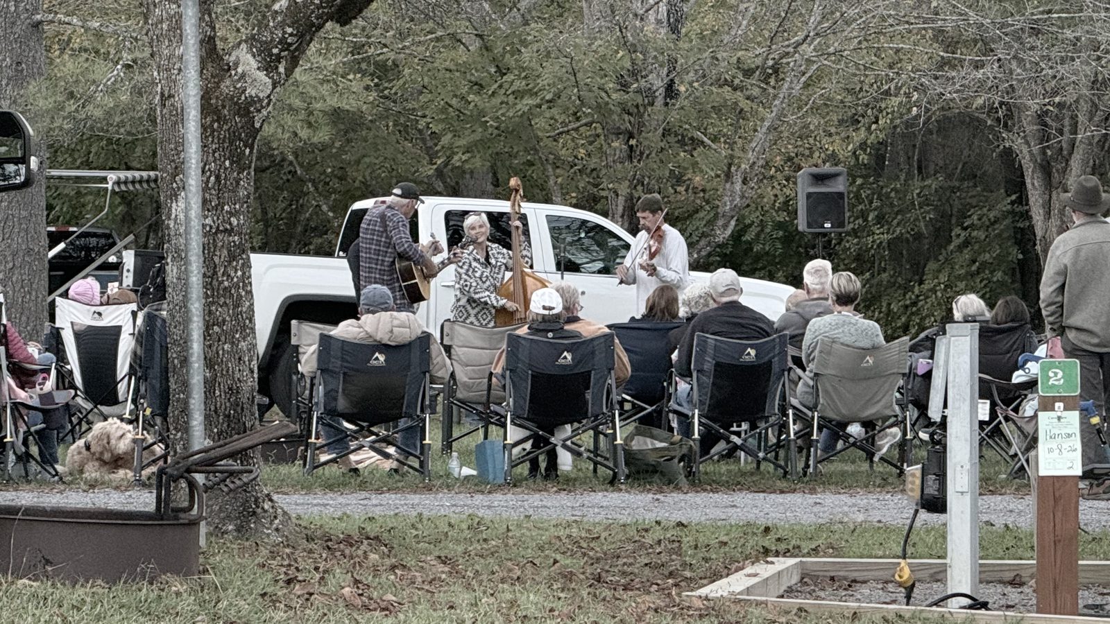 A bluegrass band playing in front of a white pickup truck at the campground while an audience sits in camp chairs on the gravel in front of them, with trees behind