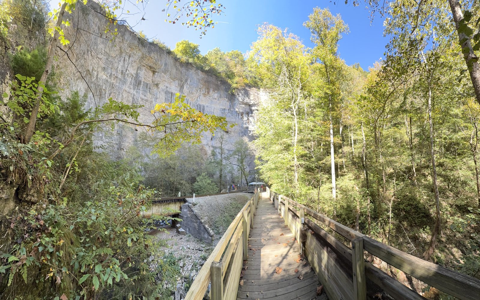 A wooden boardwalk running beside a gravel railbed below a tall limestone cliff, with autumn trees along the right side