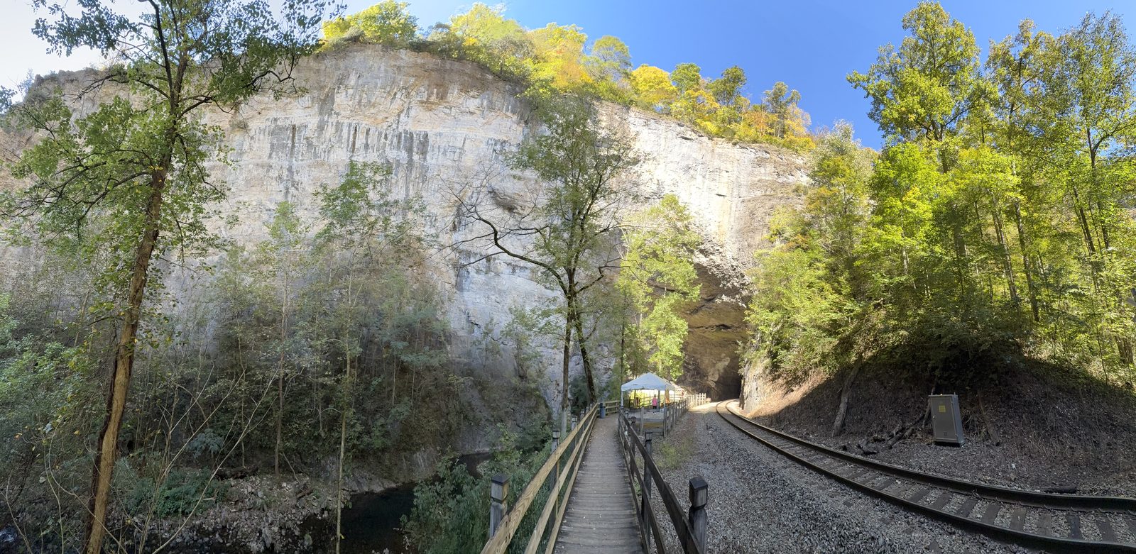 The boardwalk and railroad tracks below a pale limestone cliff, with the tunnel entrance in the shadows beneath the rock and autumn trees on either side