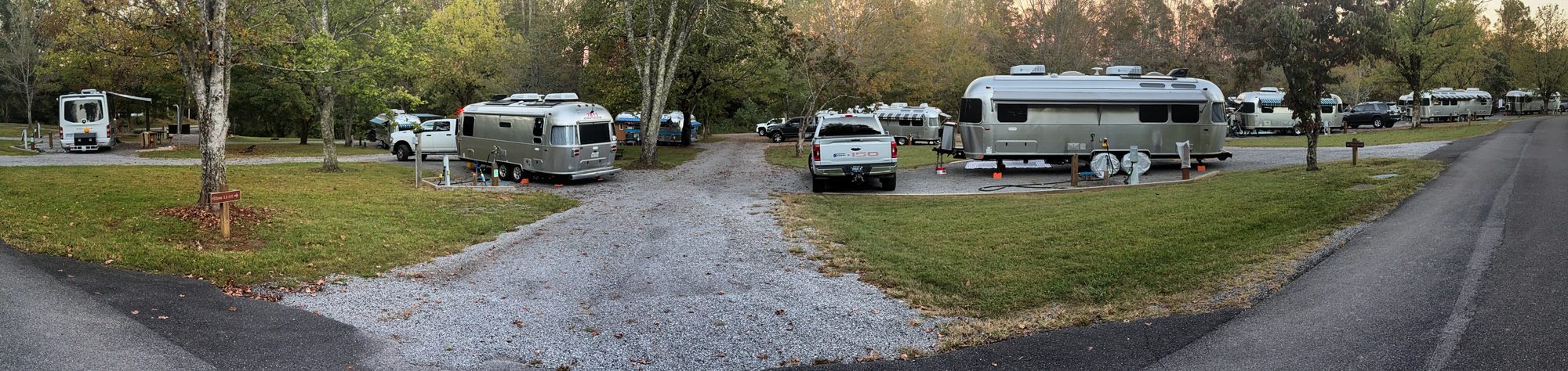 A wide view of our second base camp at golden hour: Airstream trailers and trucks parked on both sides of a curving gravel lane, with green grass, fall-colored trees and a paved road at either edge