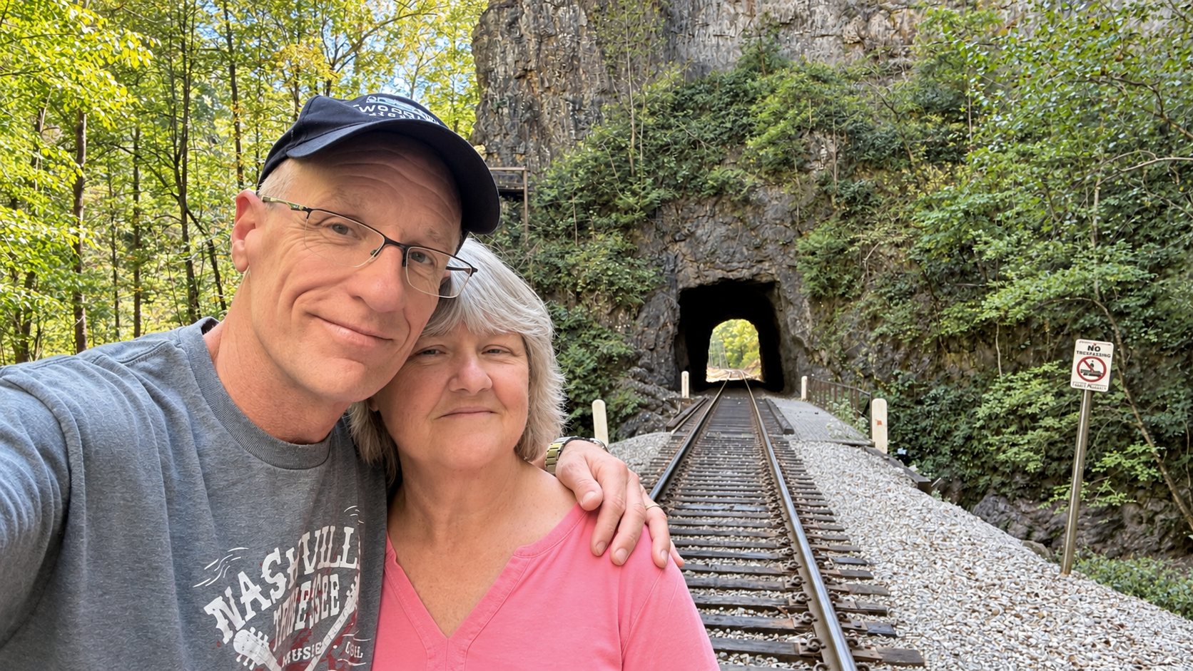 Dan in a dark cap and glasses and Heather in a pink top, smiling close together in a selfie in front of a railroad track that runs into a small tunnel under a rock cliff, with green trees and a No Trespassing sign at the right
