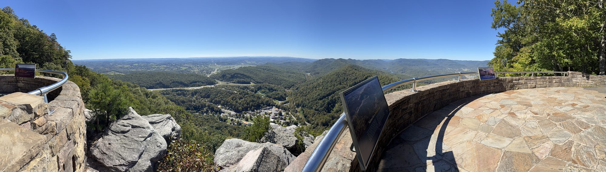 A wide view from Pinnacle Overlook over forested ridges and a valley town, with a stone retaining wall and an interpretive sign at the right edge of the viewing platform