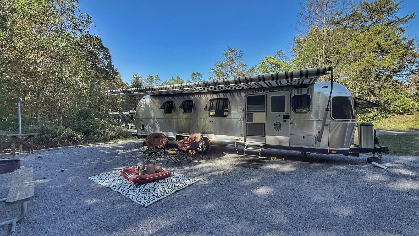 Our Airstream parked at an angle on a gravel site under trees, with the awning out, camp chairs, and a rug with a dog bed in front of it