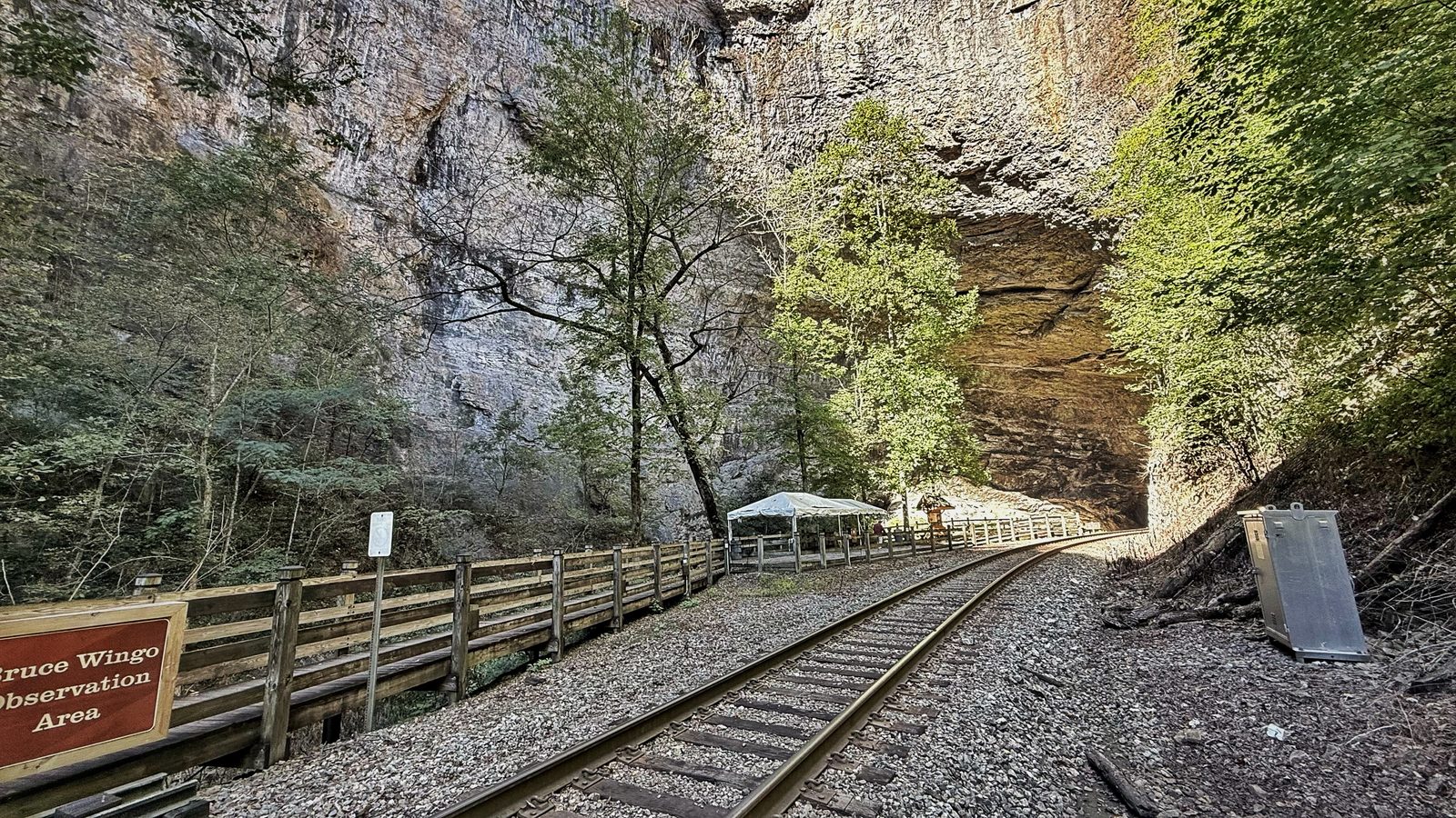 Railroad tracks curving along a limestone cliff toward the dark mouth of Natural Tunnel, with a wooden fence and a Bruce Wingo Observation Area sign on the left