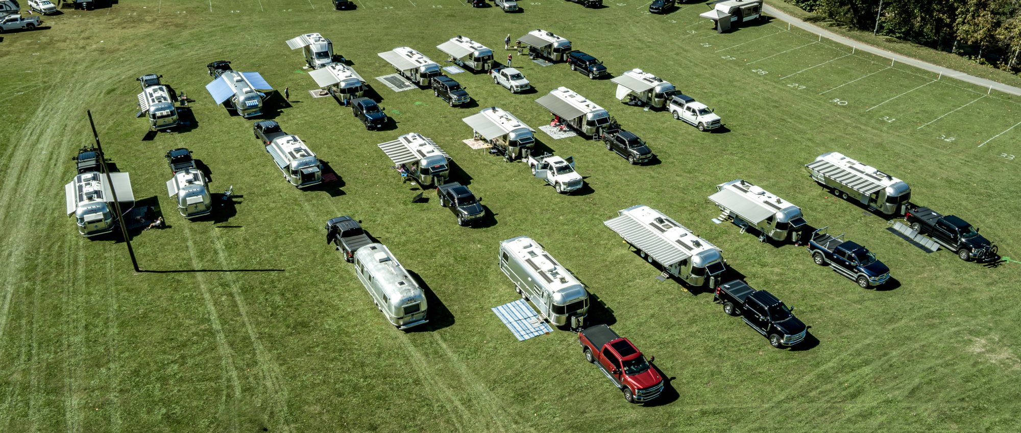 An aerial view of Airstream trailers and tow trucks parked in rows on a mowed green field, with awnings out and white yard lines on the grass at the upper right