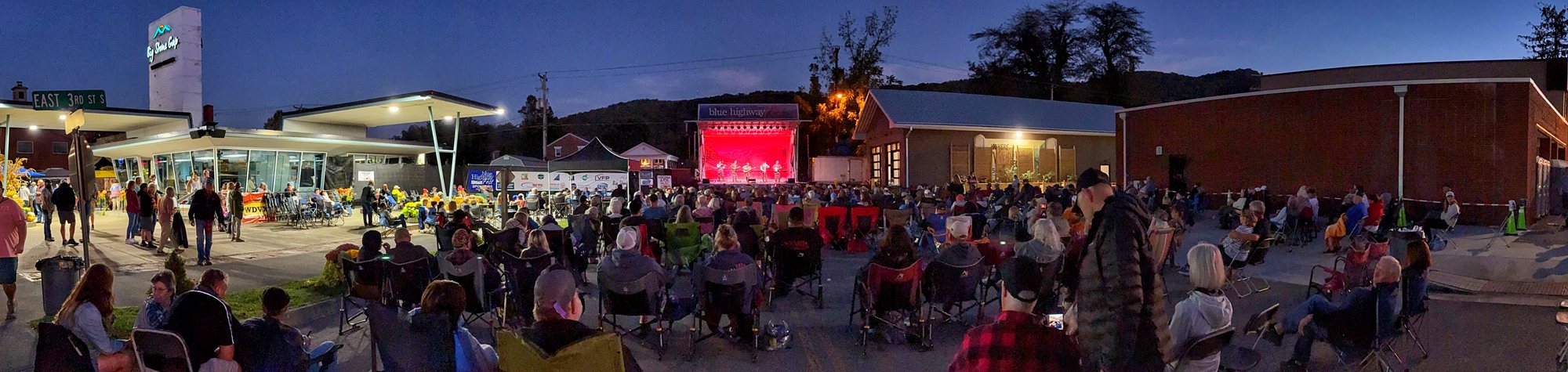 A wide night view of the festival crowd in folding chairs along East 3rd Street, facing a stage lit in red at the far end where Balsam Range is playing, with a Big Stone Gap sign tower at the left