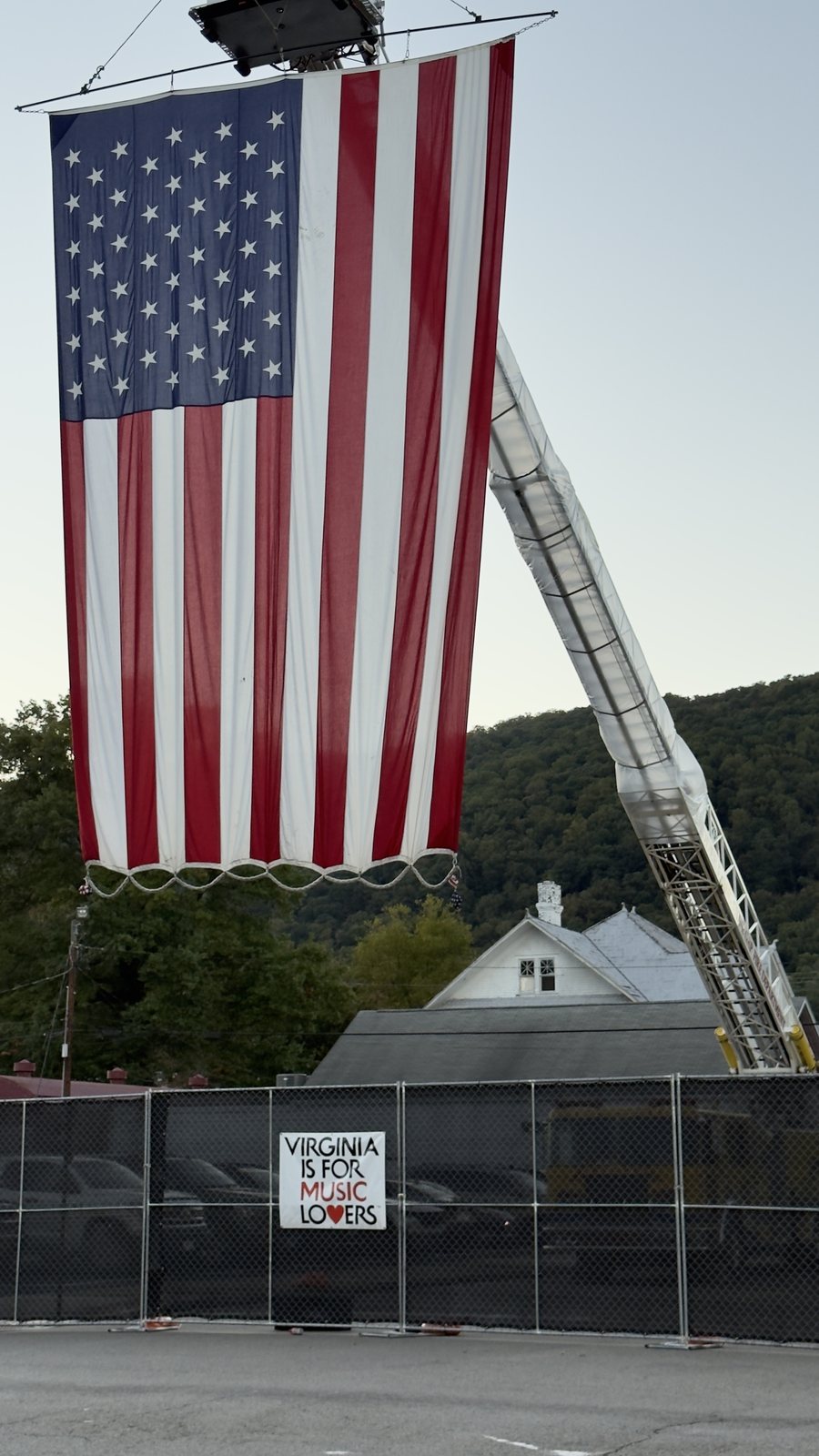 A giant American flag hanging from a raised fire truck ladder above a black fence with a sign reading Virginia is for Music Lovers