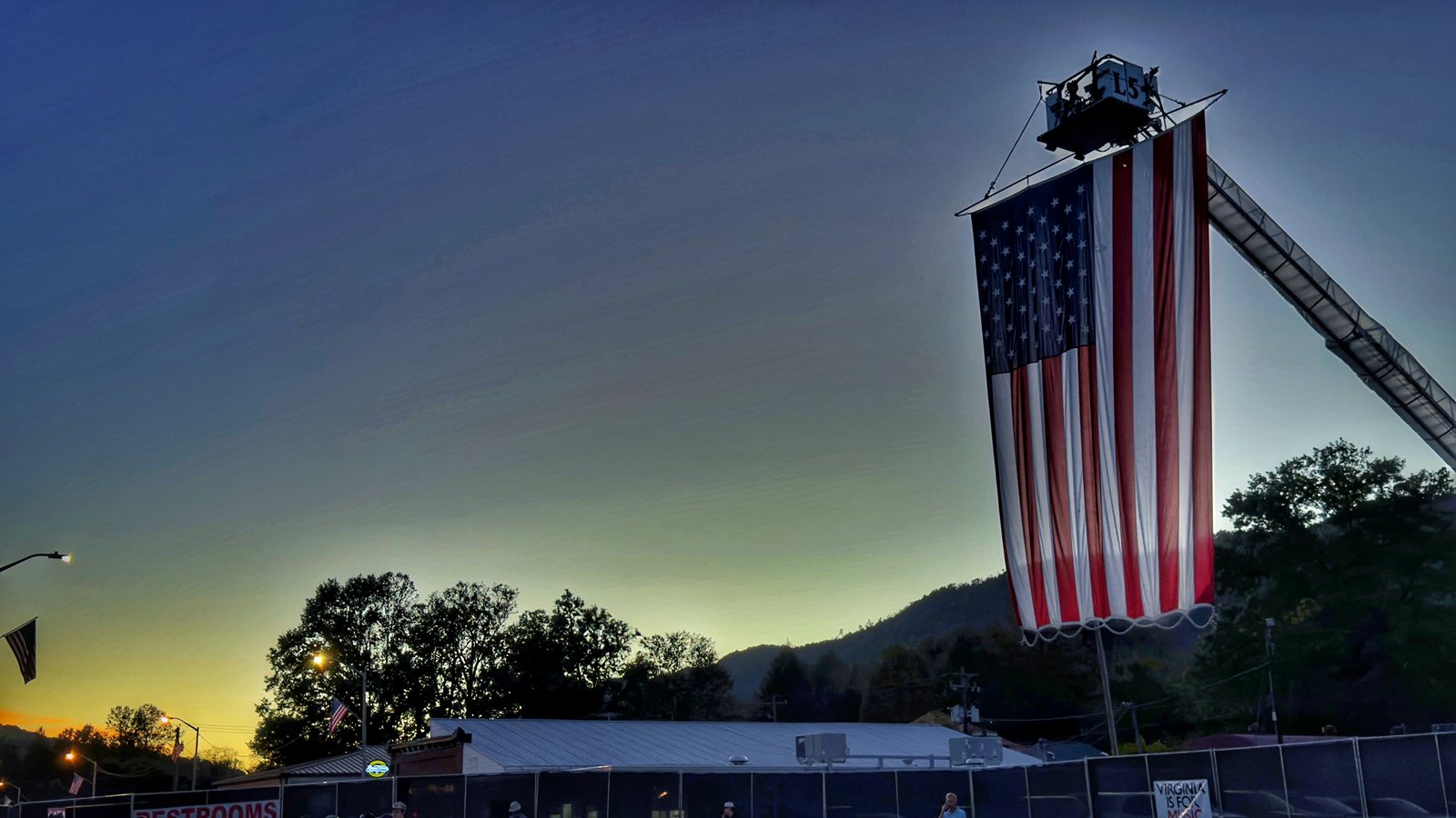 A giant American flag hanging from a raised fire truck ladder against a sunset sky, with trees, a hillside and festival fencing below