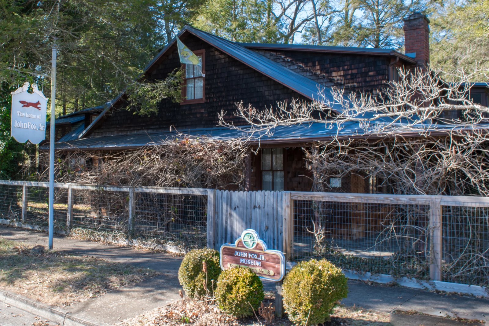The dark-shingled John Fox Jr. House Museum behind a wire and wood fence, with leafless vines across the front porch, a hanging sign reading The home of John Fox Jr. with a red fox on it, and a brown museum sign in front of two round shrubs