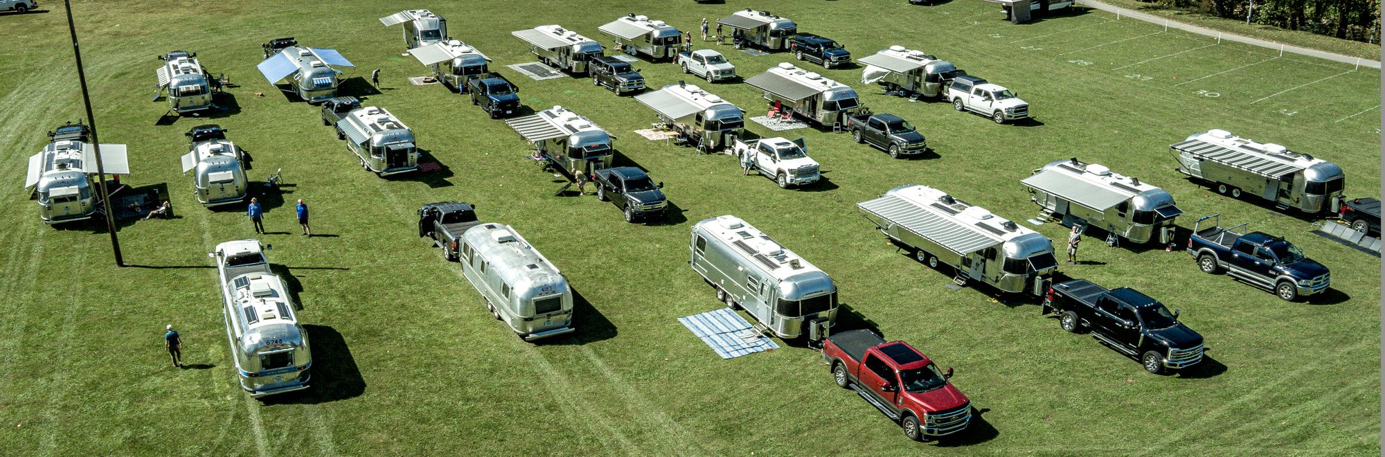 An aerial view of Airstream trailers and tow trucks parked in rows on a green grass field, with awnings out and white yard lines marked on the grass at the upper right