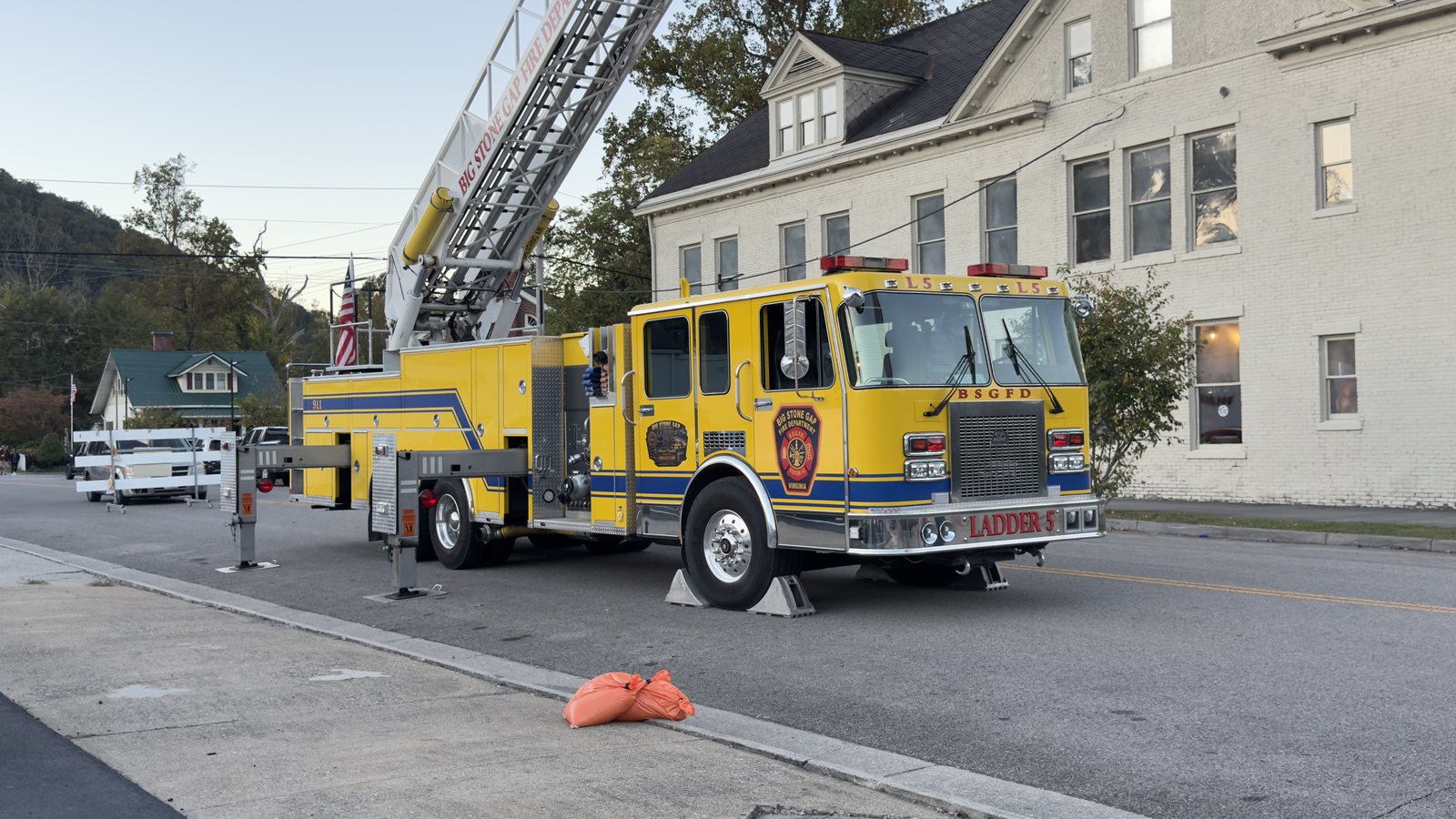 A yellow Big Stone Gap Fire Department ladder truck parked on a downtown street with its stabilizers down and its ladder raised, beside a large cream-colored building