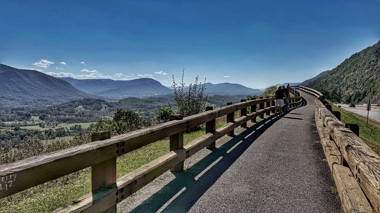 A paved path and wooden rail along the edge of the overlook, with two people walking away and distant mountains across Powell Valley