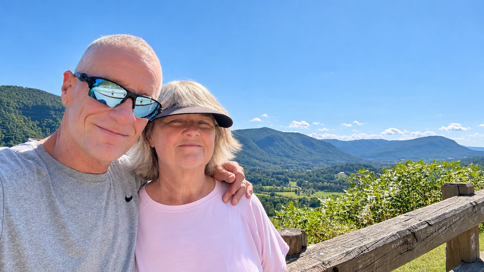 Dan in a gray shirt and sunglasses and Heather in a pink shirt and visor, smiling close together in a selfie at the overlook, with Powell Valley and mountains behind them