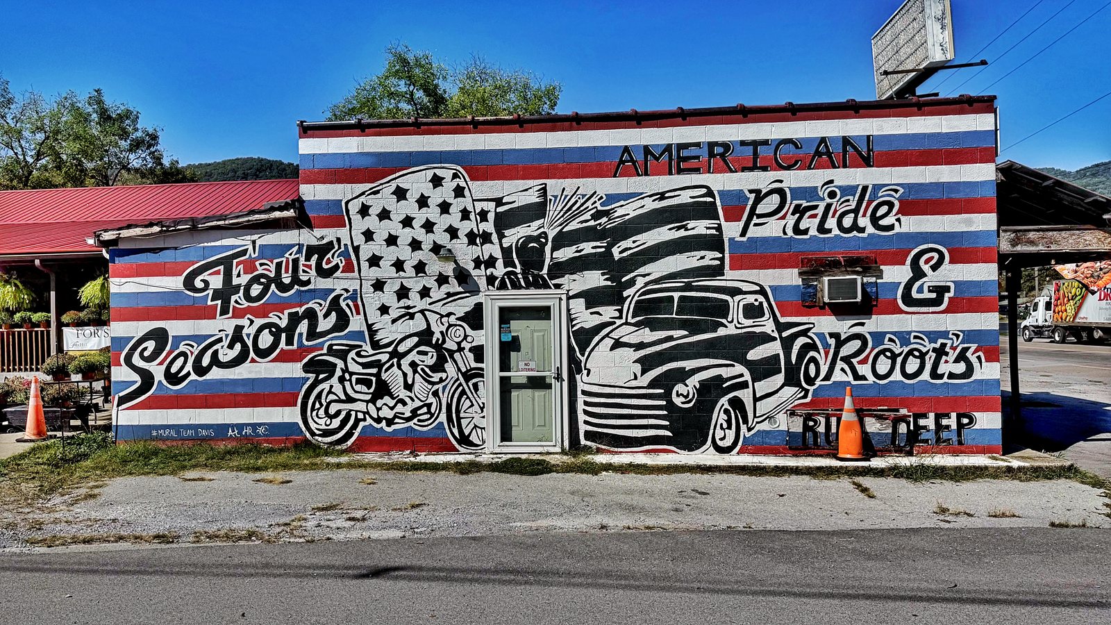 A black, white, red and blue mural on a roadside building showing an eagle and flag, a motorcycle and an old pickup truck, with the words American Pride and Roots Run Deep and Four Seasons