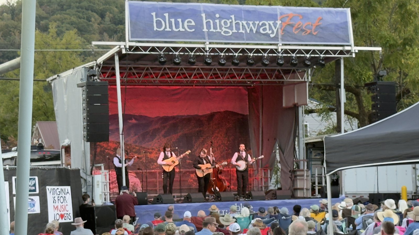 A band of five musicians on the Blue Highway Fest stage, with a mountain scene behind them and a crowd in folding chairs in the foreground