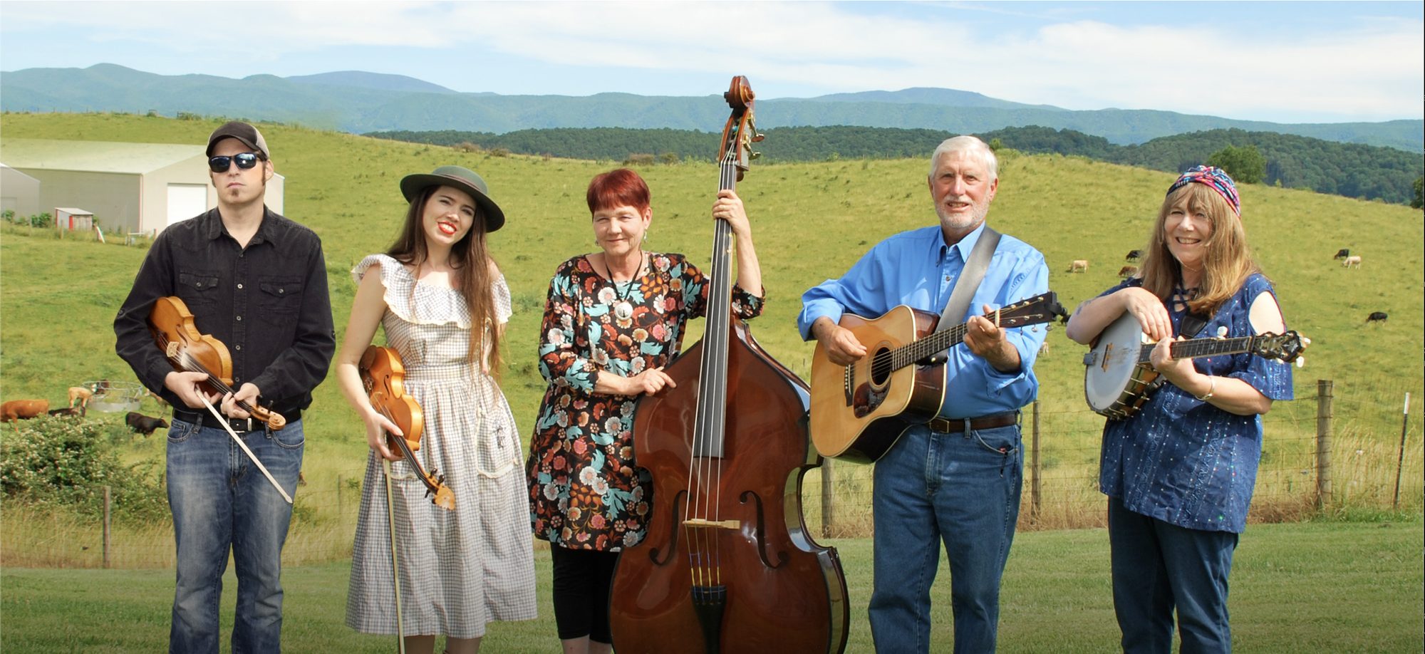 The five members of the Whitetop Mountain Band standing together in a green pasture with blue mountains behind them, holding two fiddles, an upright bass, a guitar and a banjo