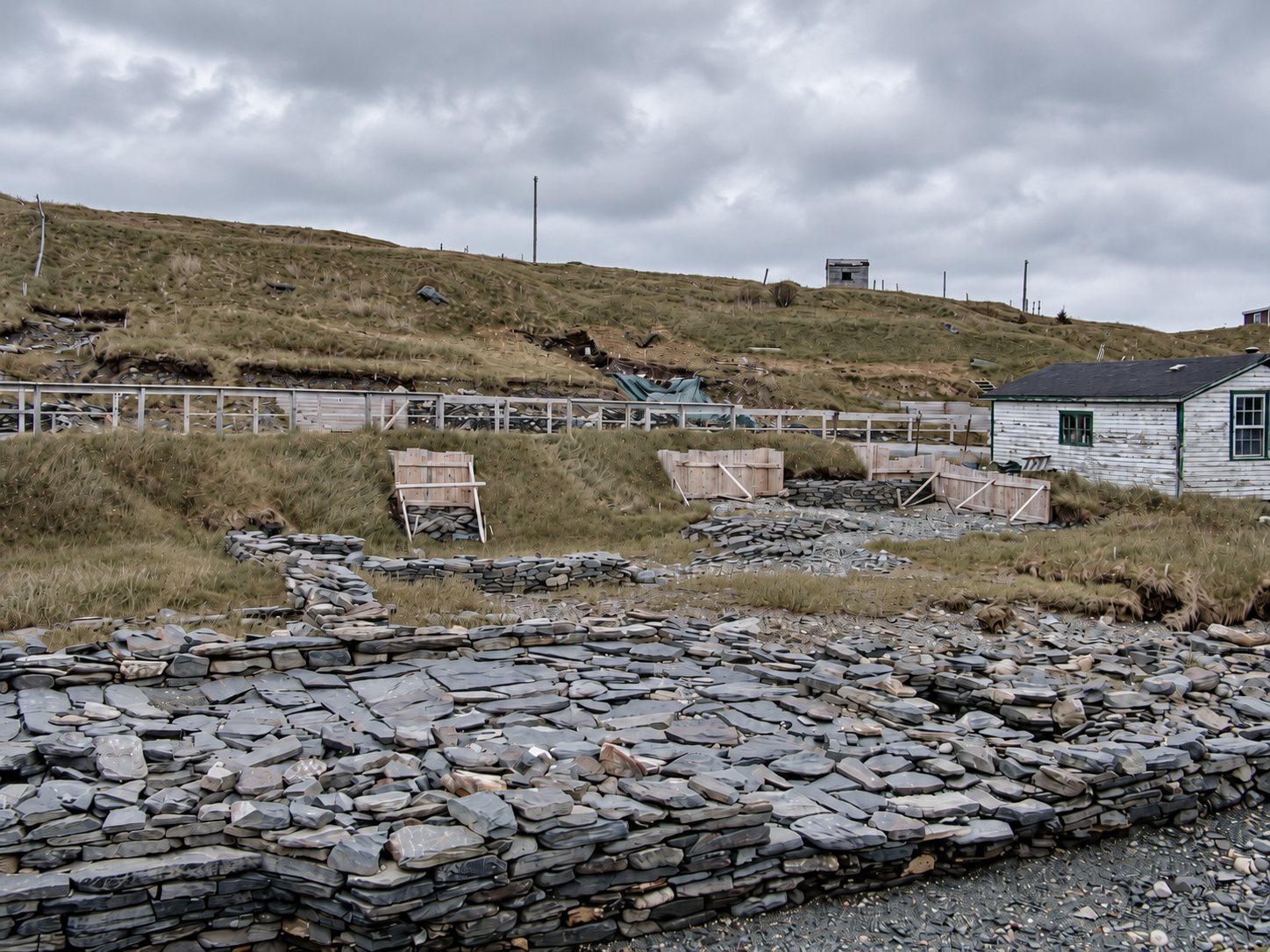 Stone foundation walls and wood-framed excavation areas below a grassy hillside at the Colony of Avalon site, with a white shed at the right