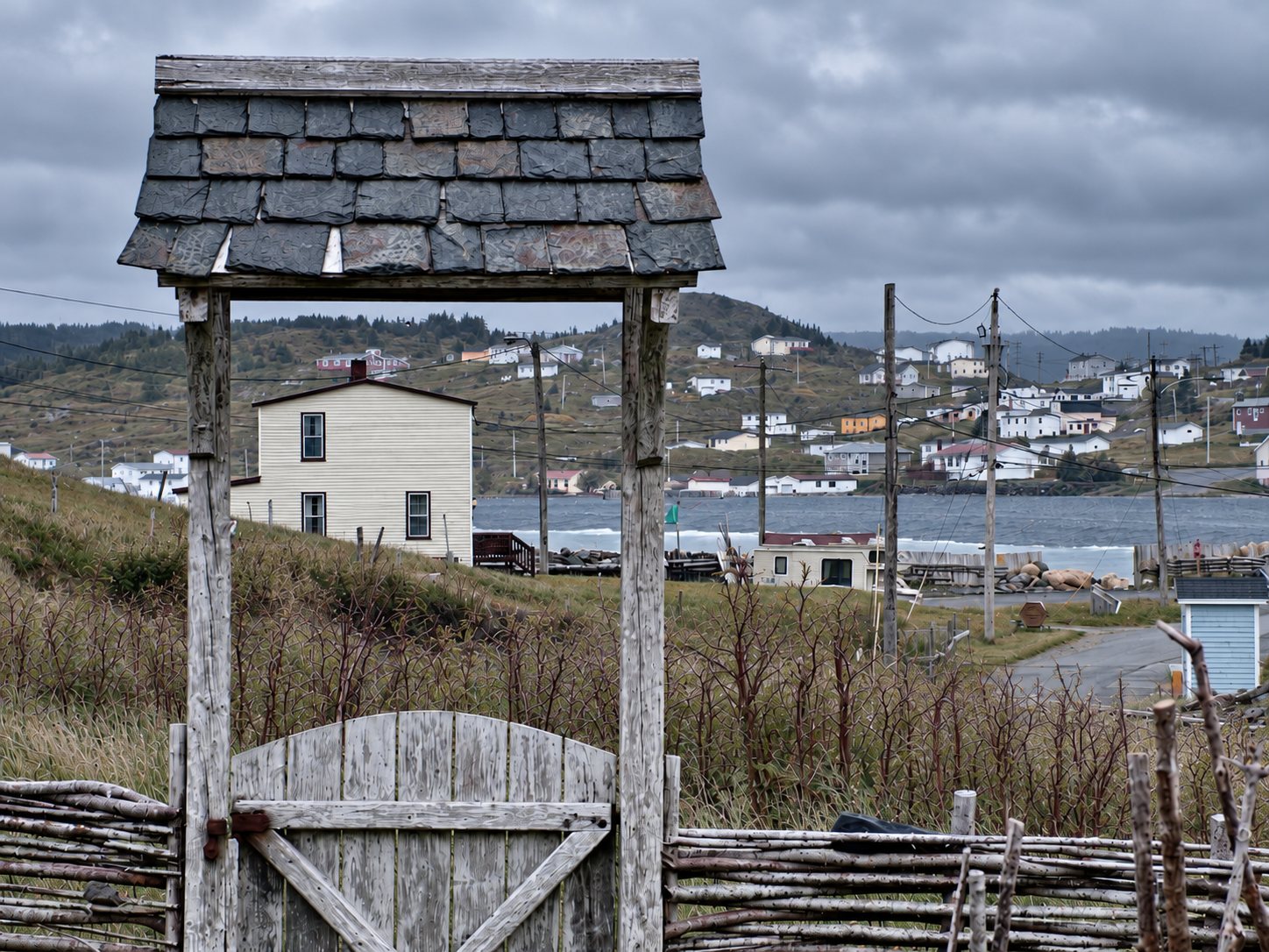 A weathered wooden gate with a slate roof and a fence of woven branches, with houses and an inlet behind it under a cloudy sky