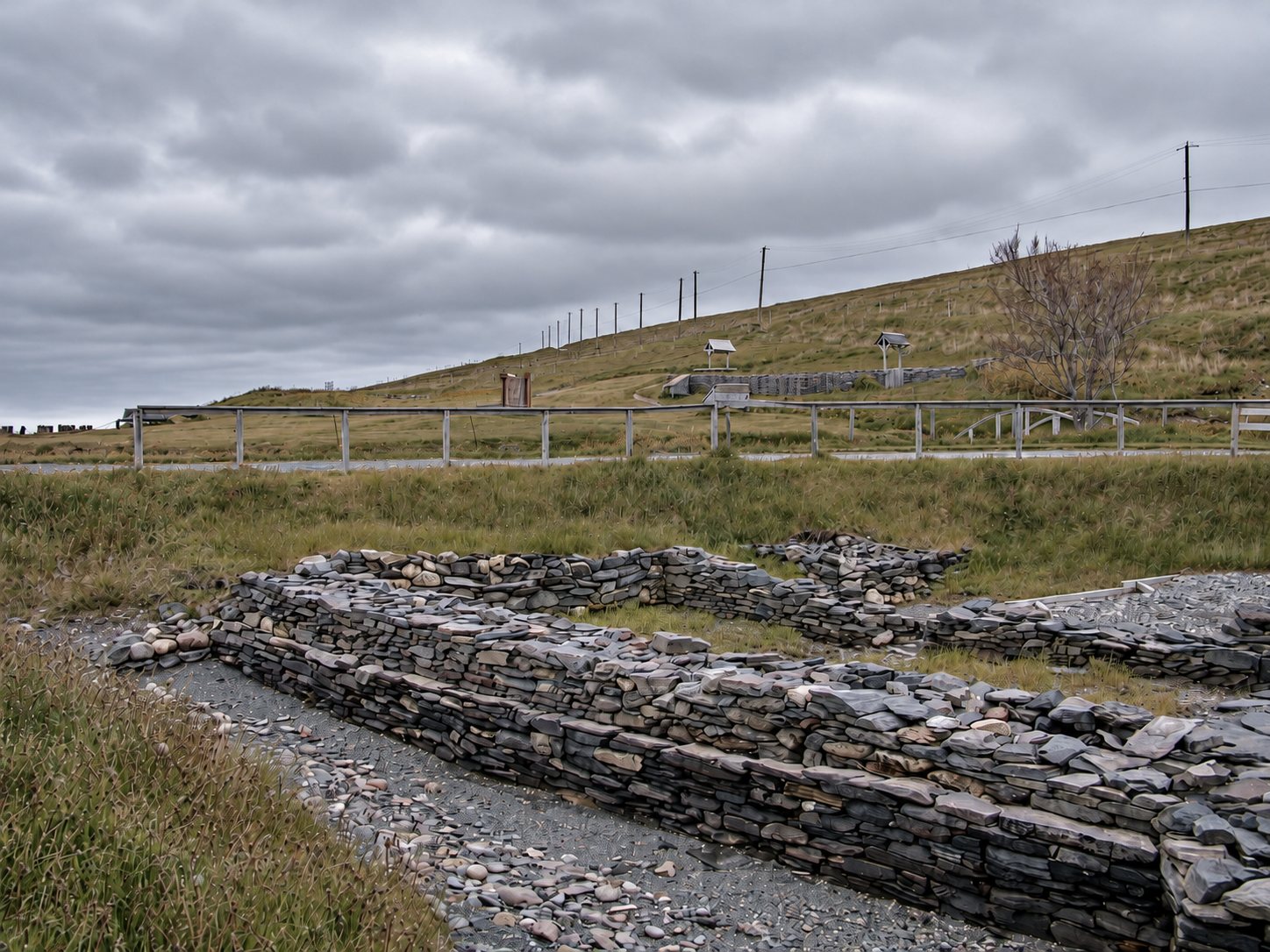 Low stone foundation walls and a pebble floor at the Colony of Avalon site, with a grassy hillside and interpretive signs behind