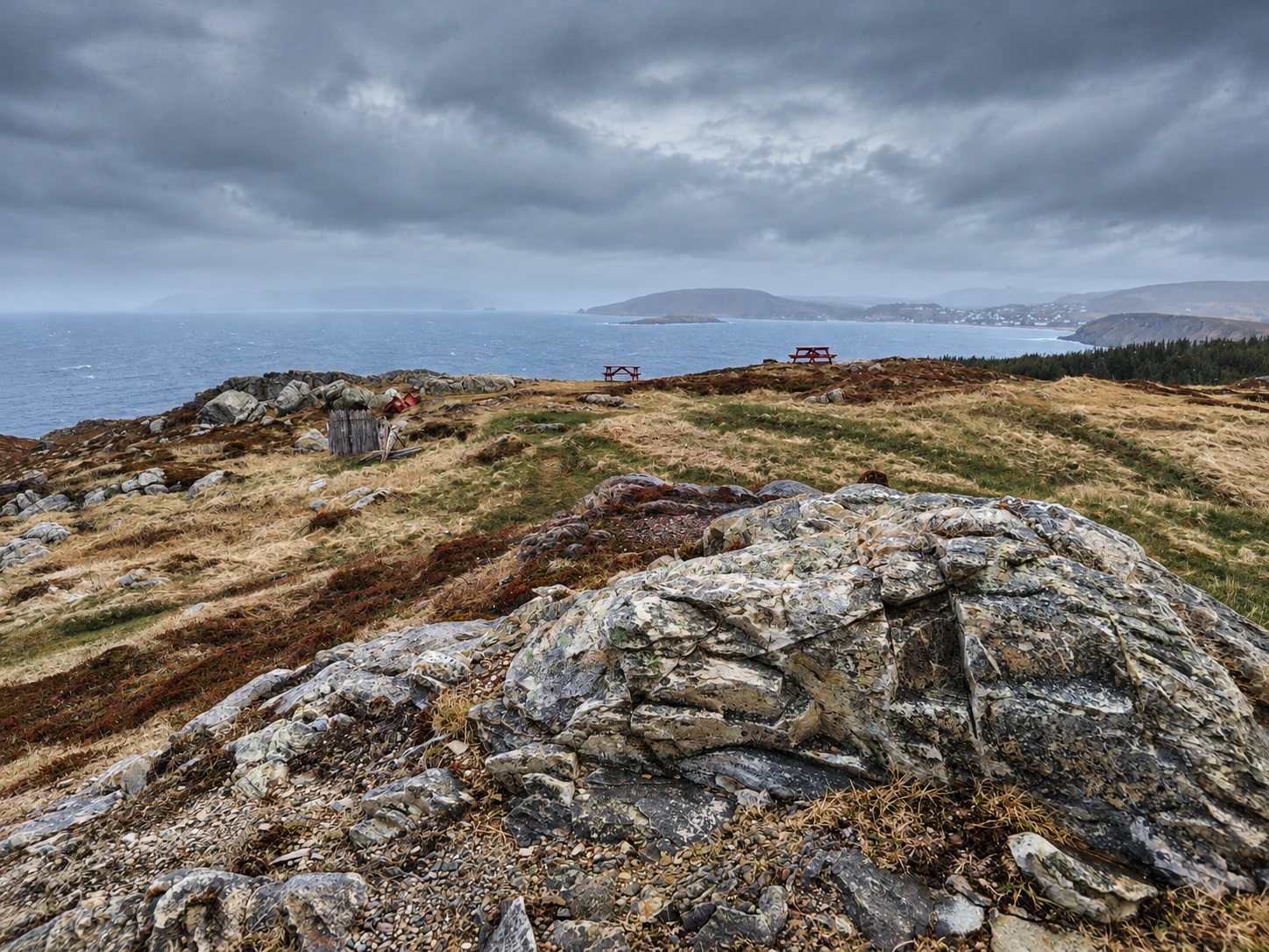 Looking south from the lighthouse, two red picnic tables on the sloped grassy headland, with a rocky outcrop in the foreground and the coast beyond