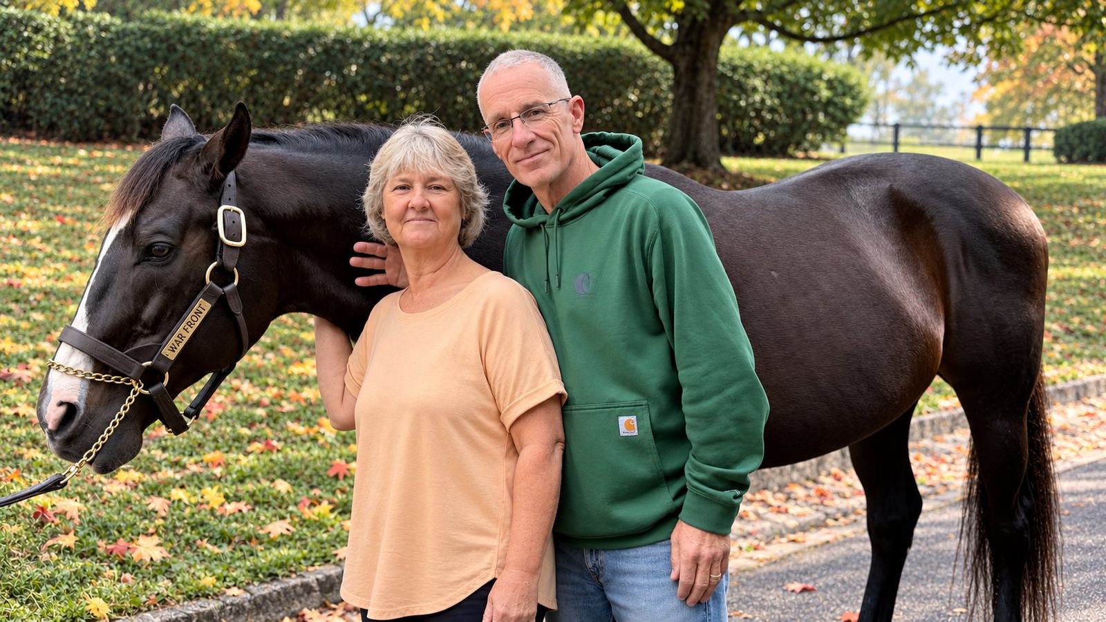 Heather in a peach top and Dan in a green hoodie beside the dark bay stallion War Front, whose halter nameplate reads War Front, on a leafy lawn