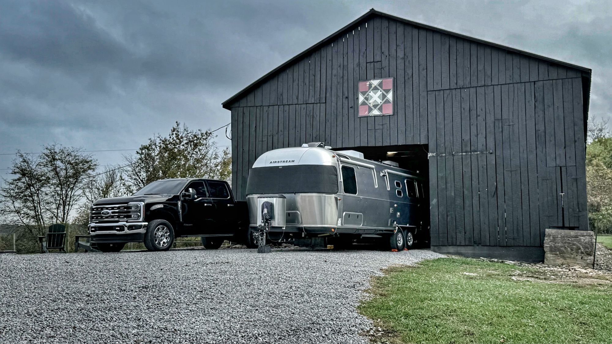 Our black truck and silver Airstream parked on gravel, with the Airstream halfway inside a black barn with a quilt-block design, under gray clouds