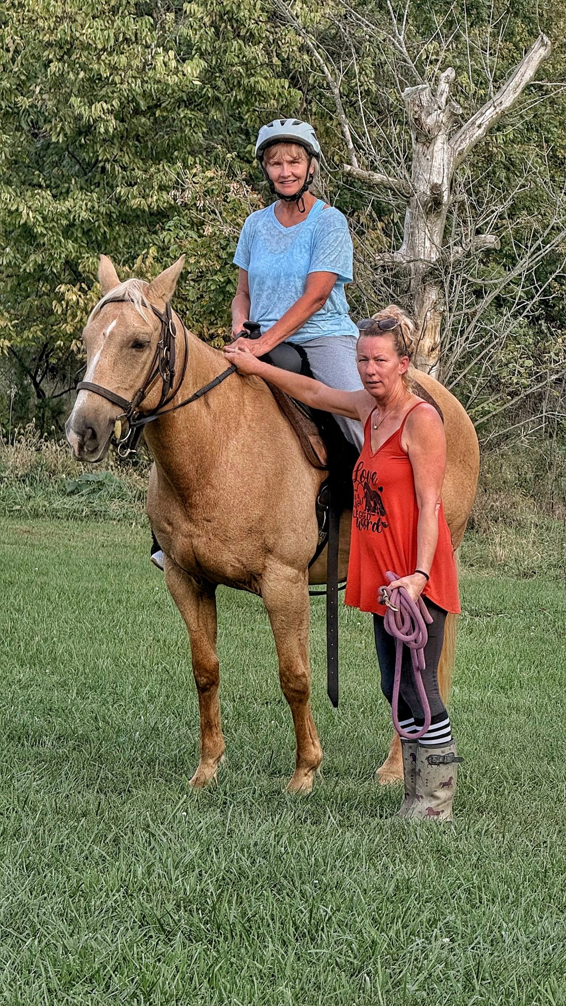 A rider in a light blue shirt and white helmet on a palomino horse in a grassy field, with Brock in a red top standing at the horse's head