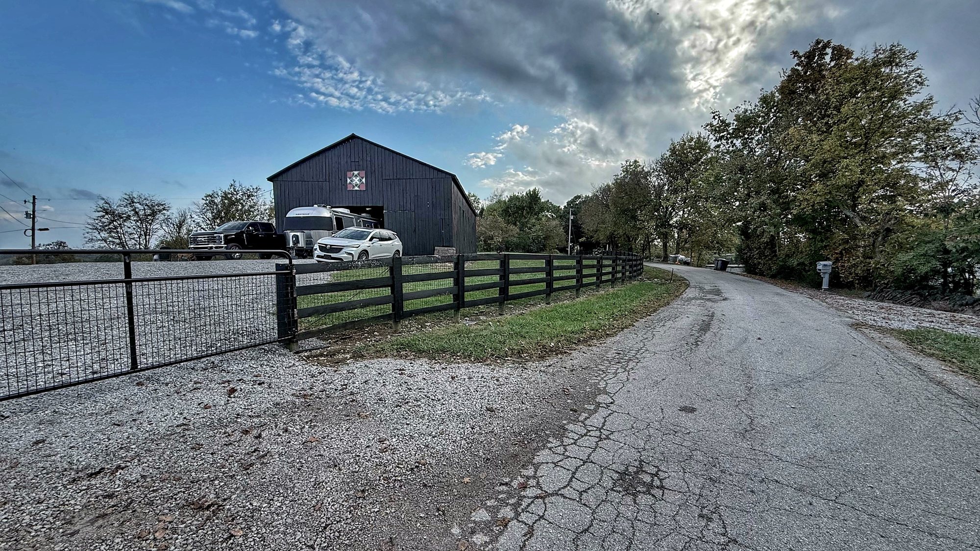 A narrow, cracked country road curving past a black fence toward a black barn, with our truck and Airstream parked near it under a blue sky
