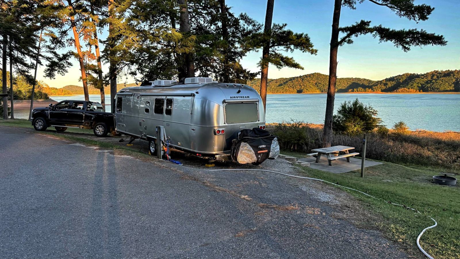 Our black truck and silver Airstream parked on a gravel site beside Norris Lake, under pine trees with a picnic table and forested hills beyond