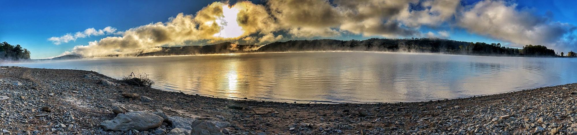 Morning mist rising off Norris Lake under glowing clouds, with a rocky shoreline in the foreground