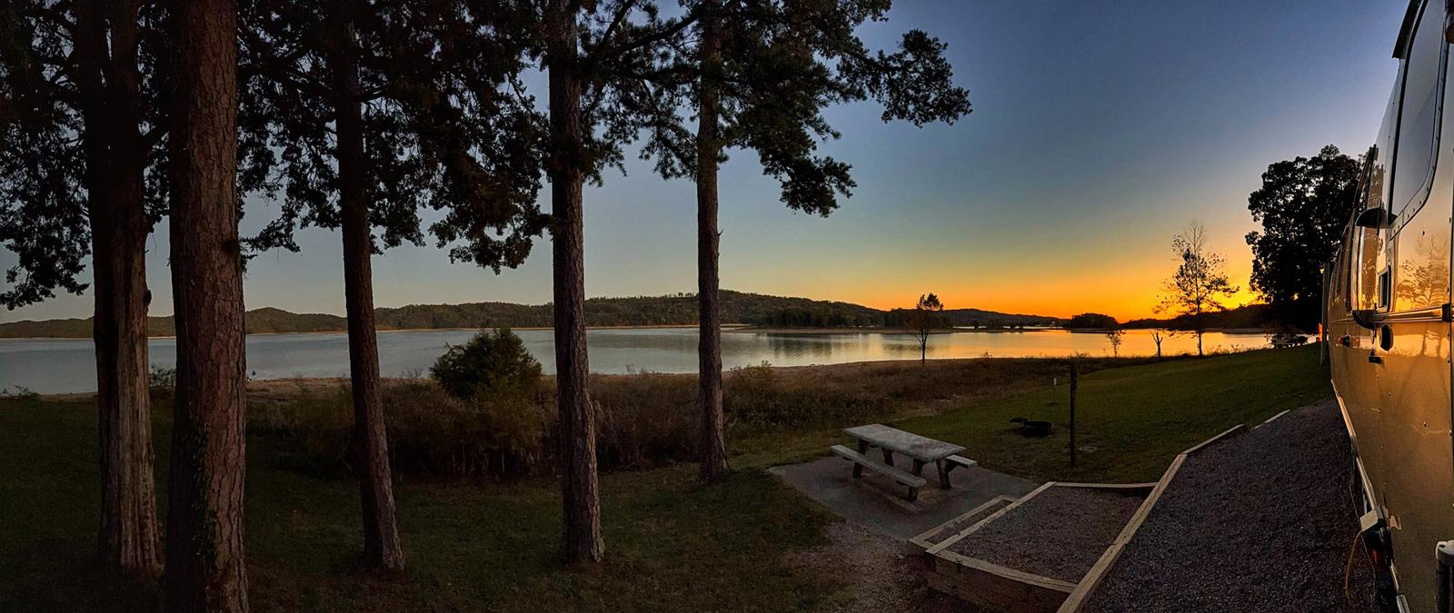Sunset over Norris Lake with an orange glow behind dark hills and trees, our Airstream at the right and a picnic table in the foreground