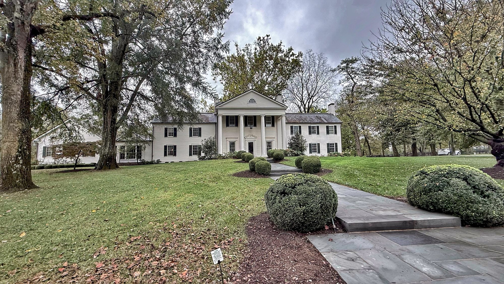 A white-columned house set back among tall old trees on a lawn, with a stone path and clipped shrubs in the foreground under a gray sky