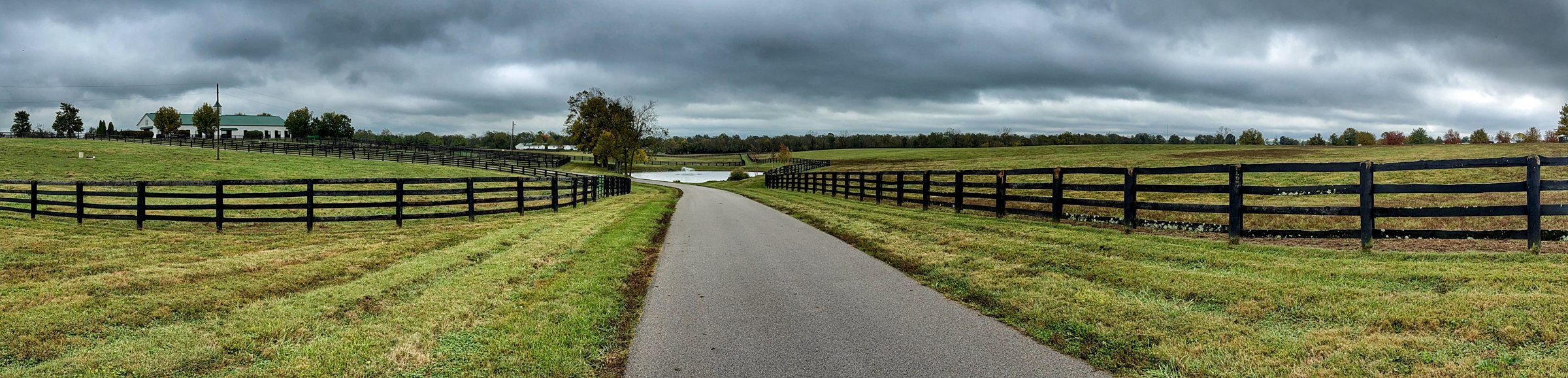 A wide view of a paved farm lane running between black board fences across green pasture under heavy gray clouds, with barns and trees in the distance