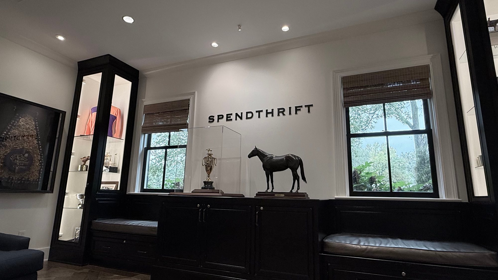 The word SPENDTHRIFT in black letters on a white wall above a cabinet holding a trophy and a bronze statue of the racehorse Authentic