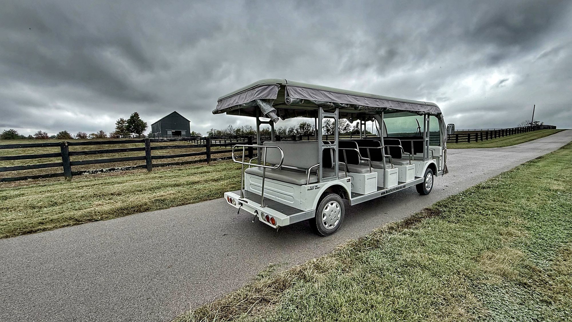 An open-sided tour vehicle with a canopy and bench seats on a paved farm lane beside a black board fence, under dark storm clouds