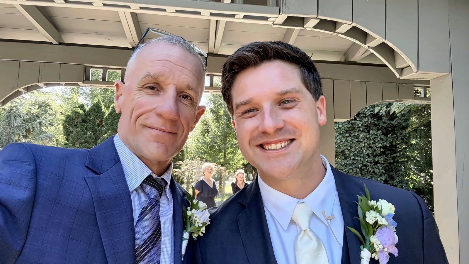 Dan and his son Ryan in navy suits with flower boutonnieres, smiling for a selfie under a covered terrace