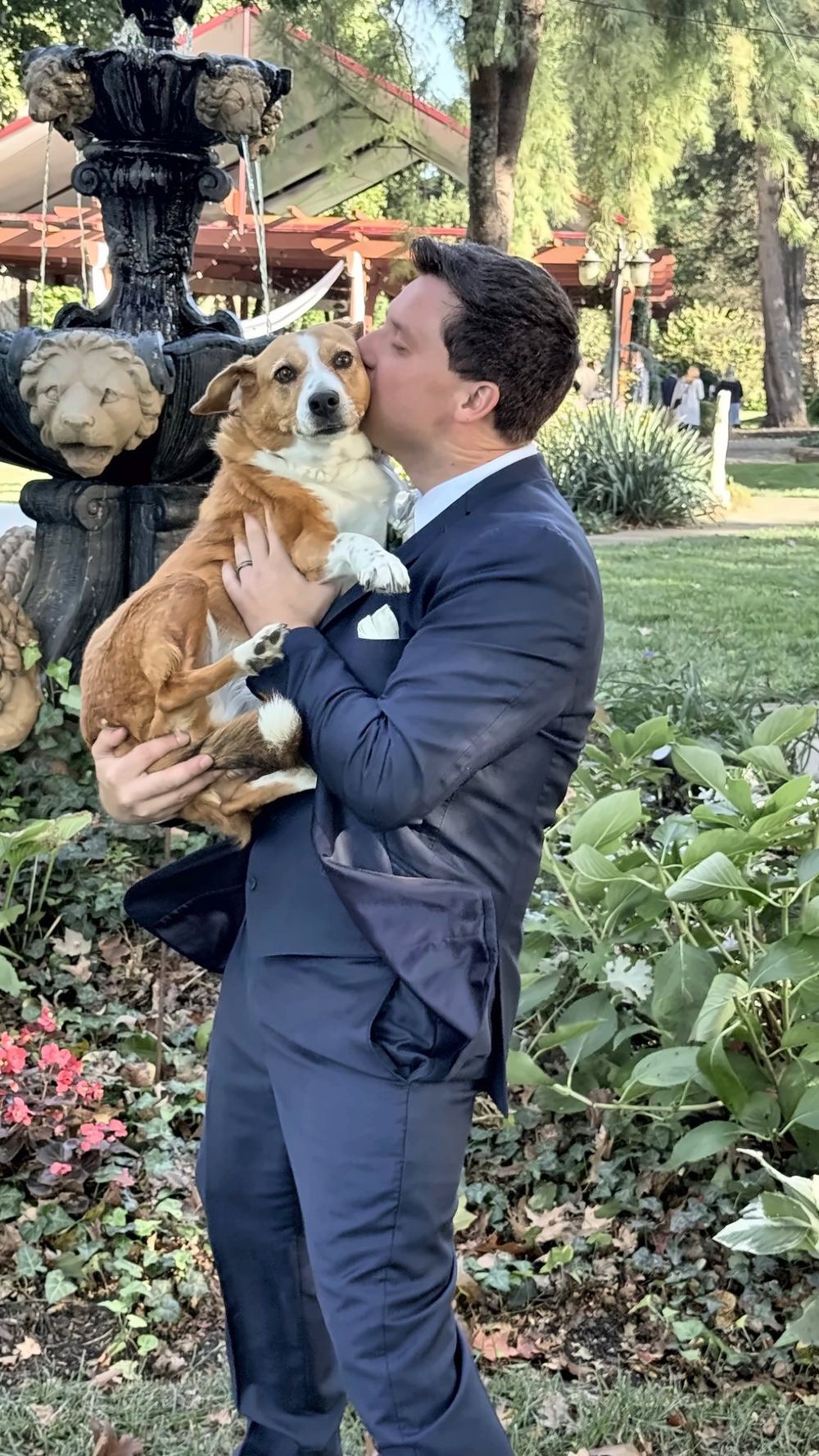 A man in a navy suit holding a corgi and kissing the dog on the head beside a stone fountain in a garden